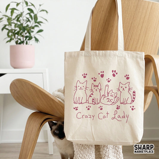 Tote bag mockup displaying a pink line art design of four cute cats and paw prints with the text Crazy Cat Lady for pet lovers and cat moms.