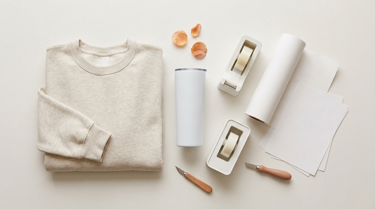 A clean workspace showing print-on-demand apparel prep with a blank sweatshirt, tumbler, and sublimation tools.