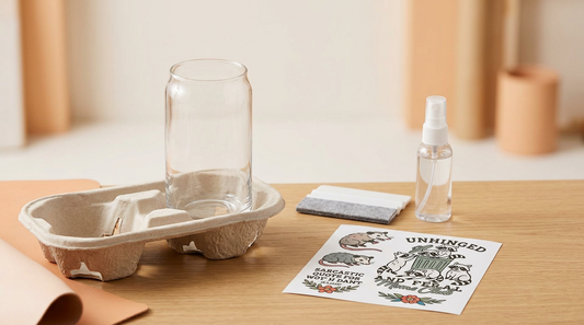 A crafter applying a UV DTF cup wrap to a clear glass can on a wooden desk surrounded by squeegees and craft supplies.