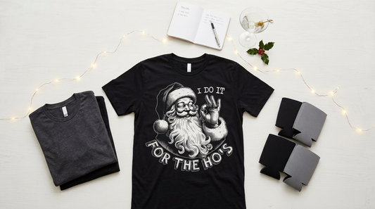 A festive workspace with blank t-shirts, can coolers, and holiday party props for creating adult humor Christmas apparel.