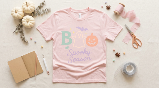 A flat lay of a light pink t-shirt blank surrounded by white mini pumpkins and crafting tools.