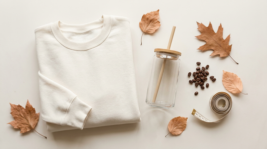 Flat lay of a cozy fall workspace with a blank crewneck sweatshirt, a glass can cup, and autumn leaves.