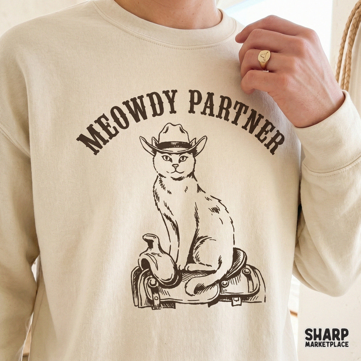 Cream colored sweatshirt featuring a brown line art design of a cat wearing a cowboy hat perched on a western saddle with Meowdy Partner text.