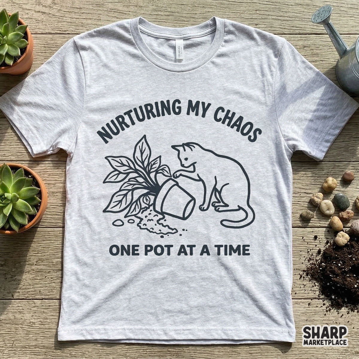A heather gray t-shirt mockup featuring a black line art graphic of a cat tipping over a houseplant with the phrase Nurturing My Chaos One Pot At A Time.