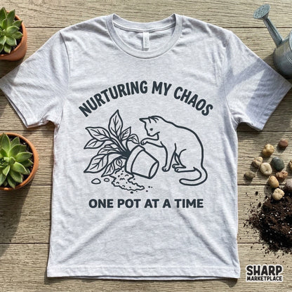 A heather gray t-shirt mockup featuring a black line art graphic of a cat tipping over a houseplant with the phrase Nurturing My Chaos One Pot At A Time.