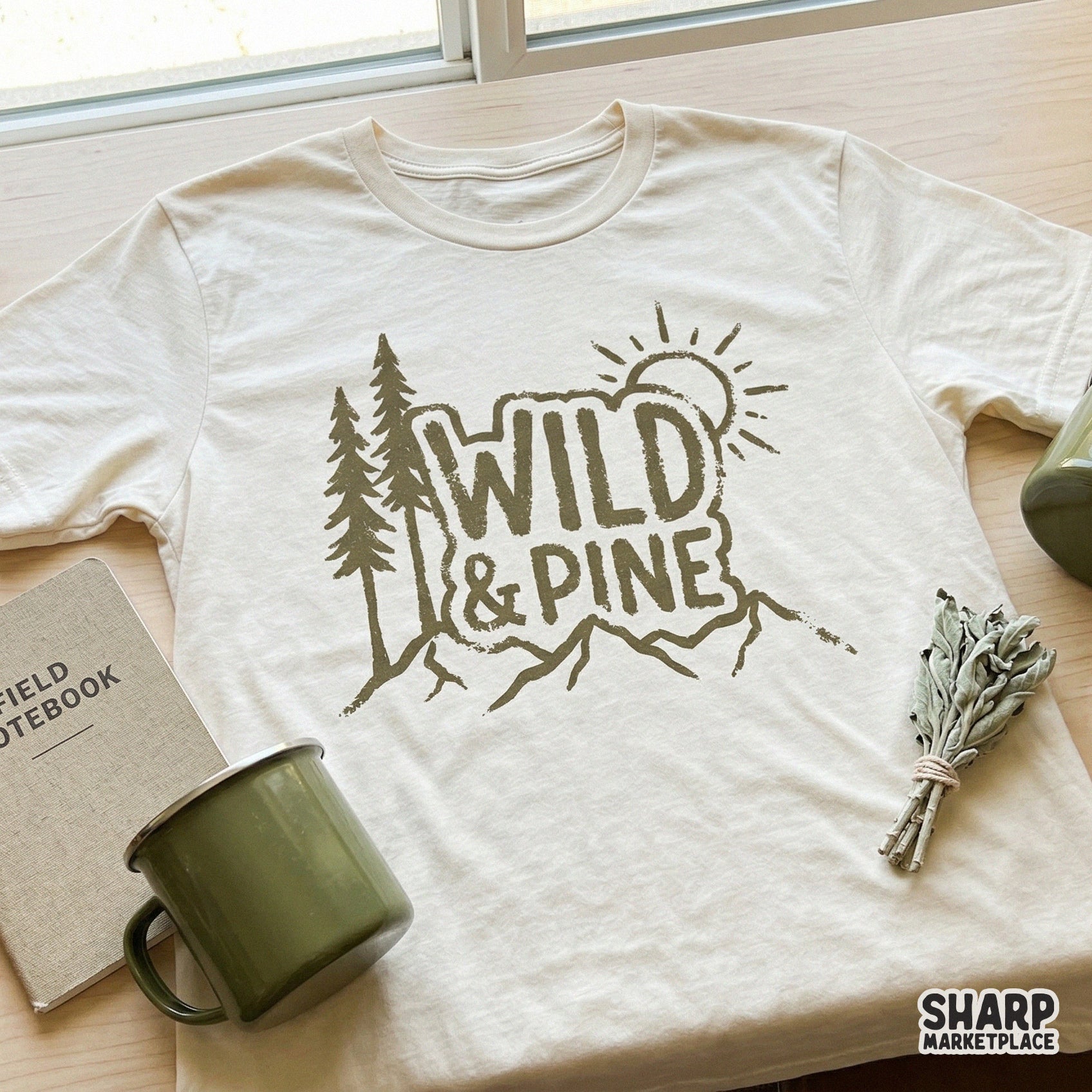 Olive green Wild and Pine digital design featuring a mountain range, stylized sun, and pine trees, shown printed on a cream colored t-shirt.