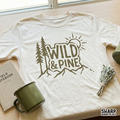 Olive green Wild and Pine digital design featuring a mountain range, stylized sun, and pine trees, shown printed on a cream colored t-shirt.
