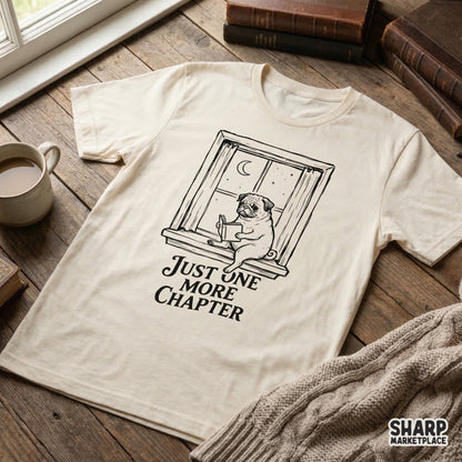 A cream colored t-shirt featuring a black ink design of a pug reading a book on a window ledge with the quote Just One More Chapter underneath.