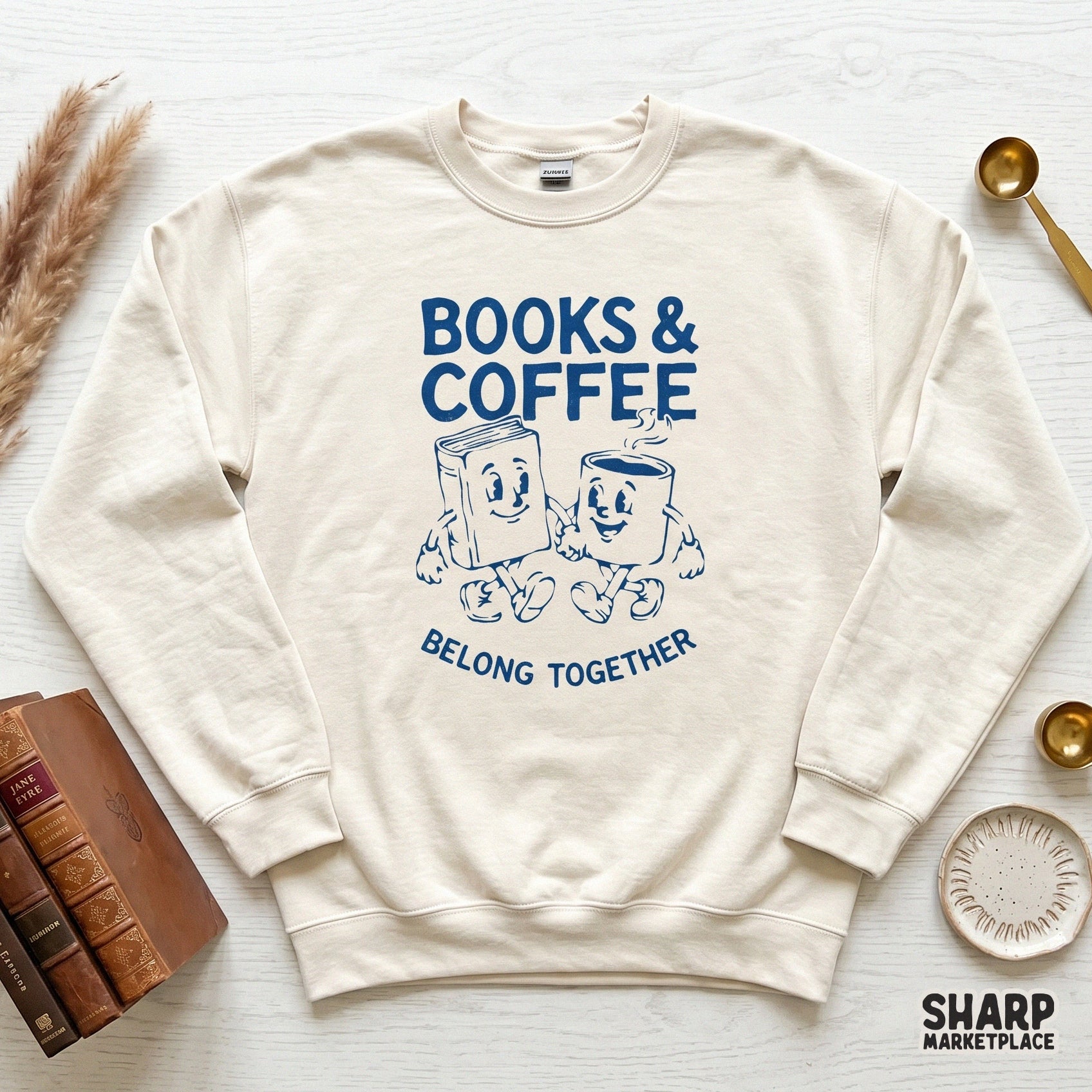 Cream-colored sweatshirt mockup featuring a vintage-style blue illustration of a happy book and coffee mug with the text Books and Coffee Belong Together.