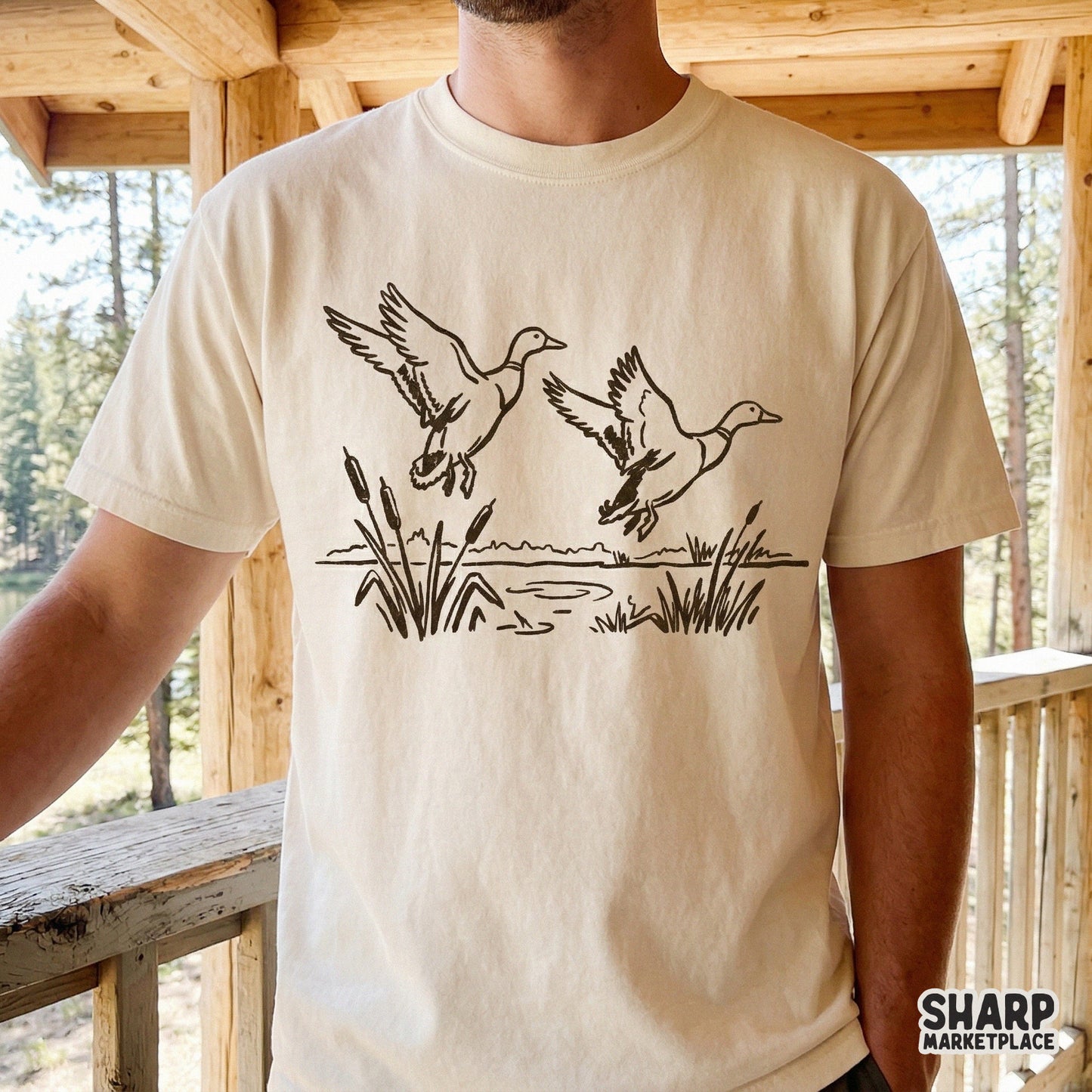 Minimalist black line art of a pair of mallards in flight over a lake, displayed on a beige t-shirt mockup for hunting and outdoor apparel.