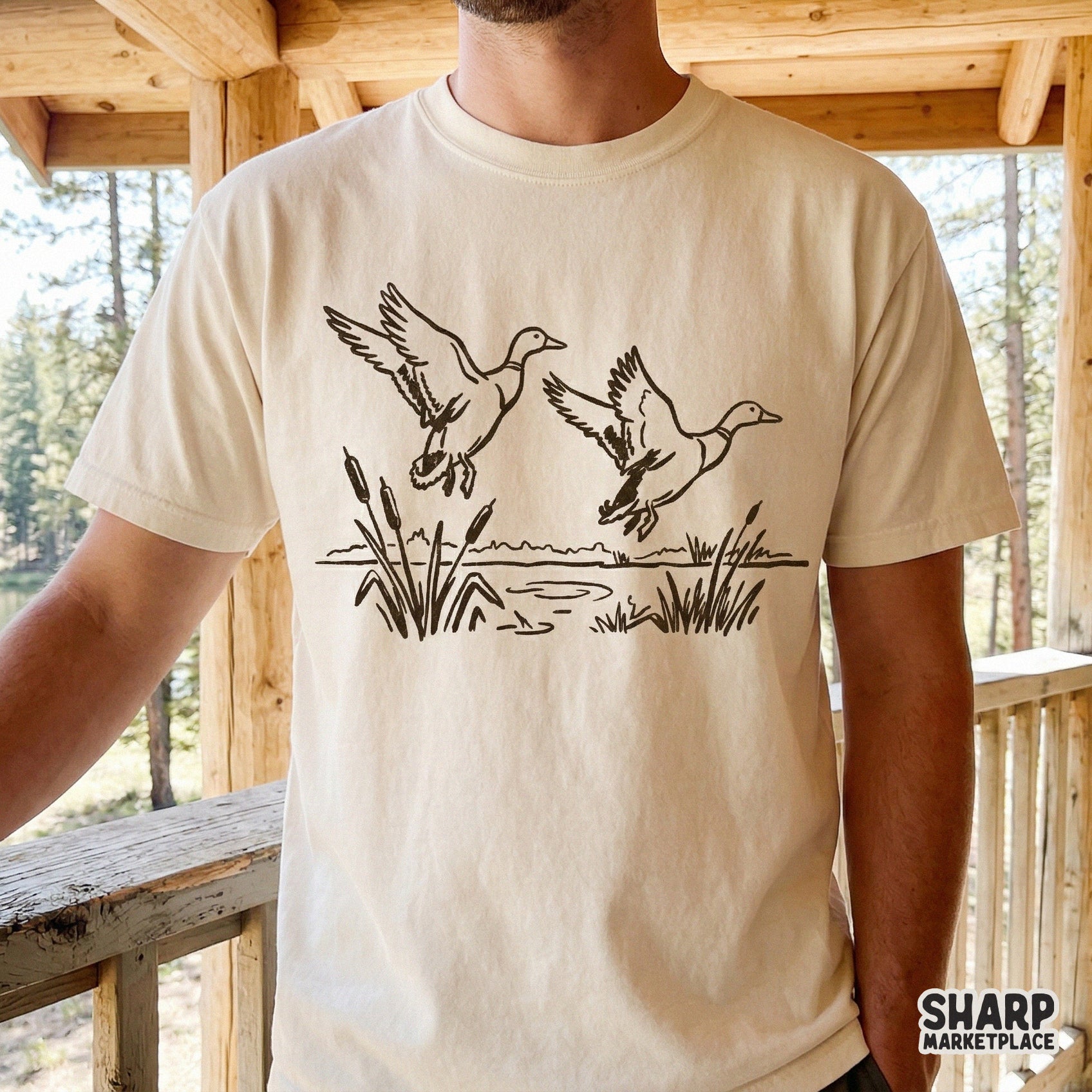 Minimalist black line art of a pair of mallards in flight over a lake, displayed on a beige t-shirt mockup for hunting and outdoor apparel.