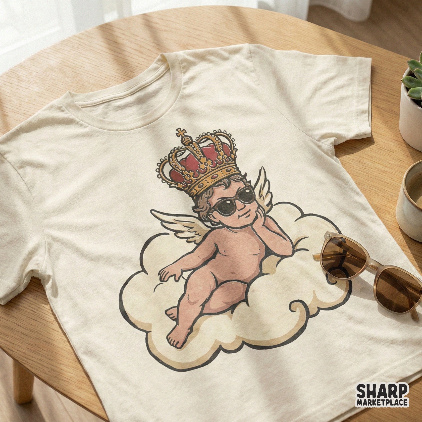 Digital design of a sassy winged baby angel with sunglasses and a crown, displayed on a cream colored t-shirt mockup.