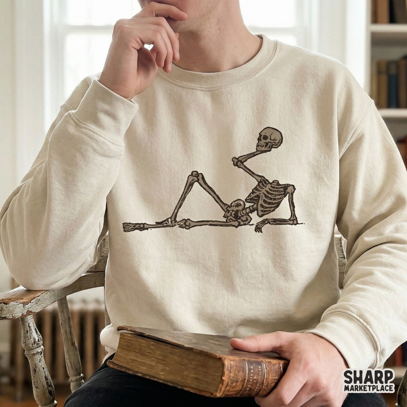 A trendy dark humor skeleton design in a sepia-toned woodcut style, shown as a graphic print on a cream-colored sweatshirt for Halloween apparel.
