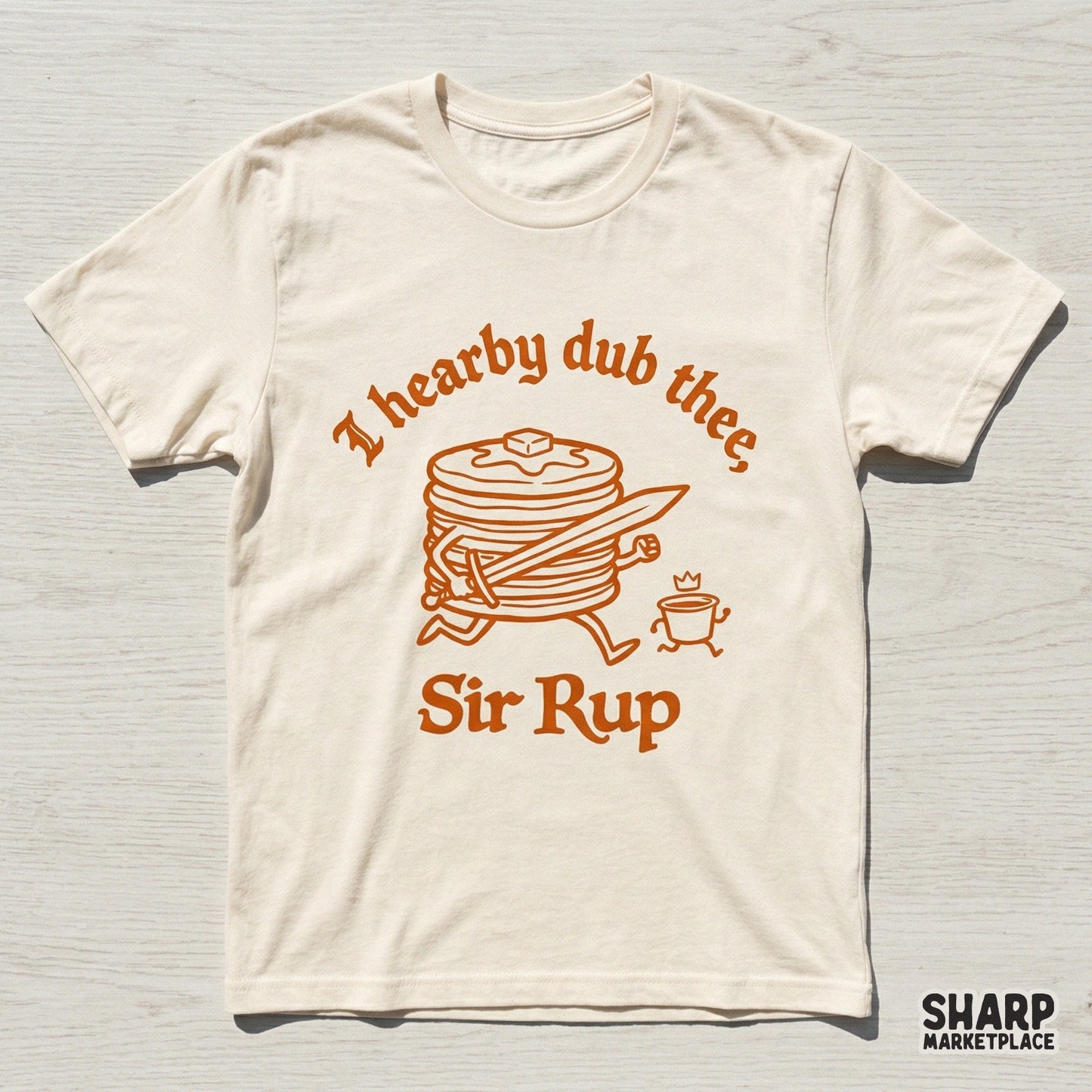 A cream-colored t-shirt mockup featuring a vintage-style orange graphic of a pancake knight with a sword and the punny quote I hearby dub thee, Sir Rup.