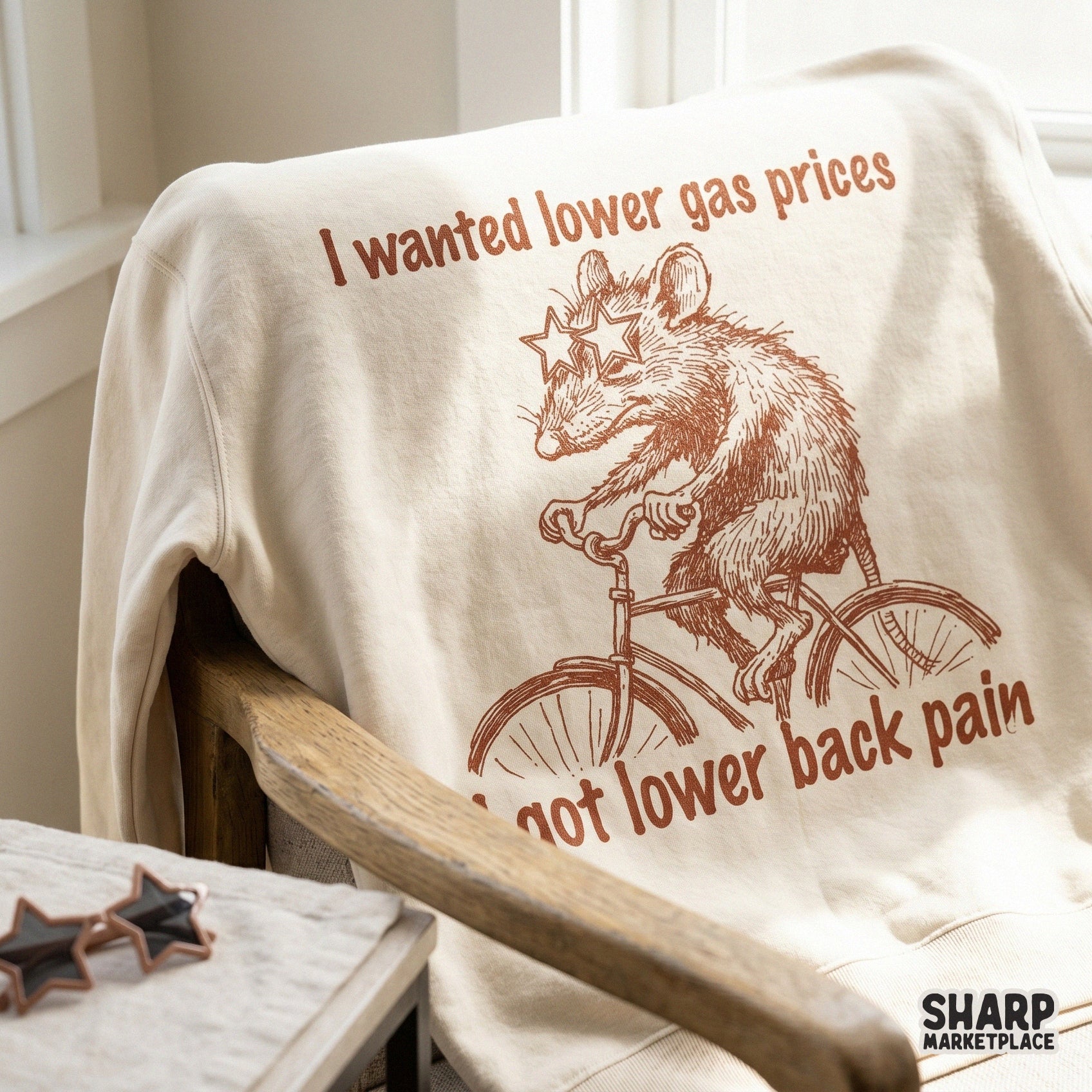 A cream-colored sweatshirt featuring a vintage-style brown illustration of a possum on a bike with the sarcastic quote about gas prices and back pain.