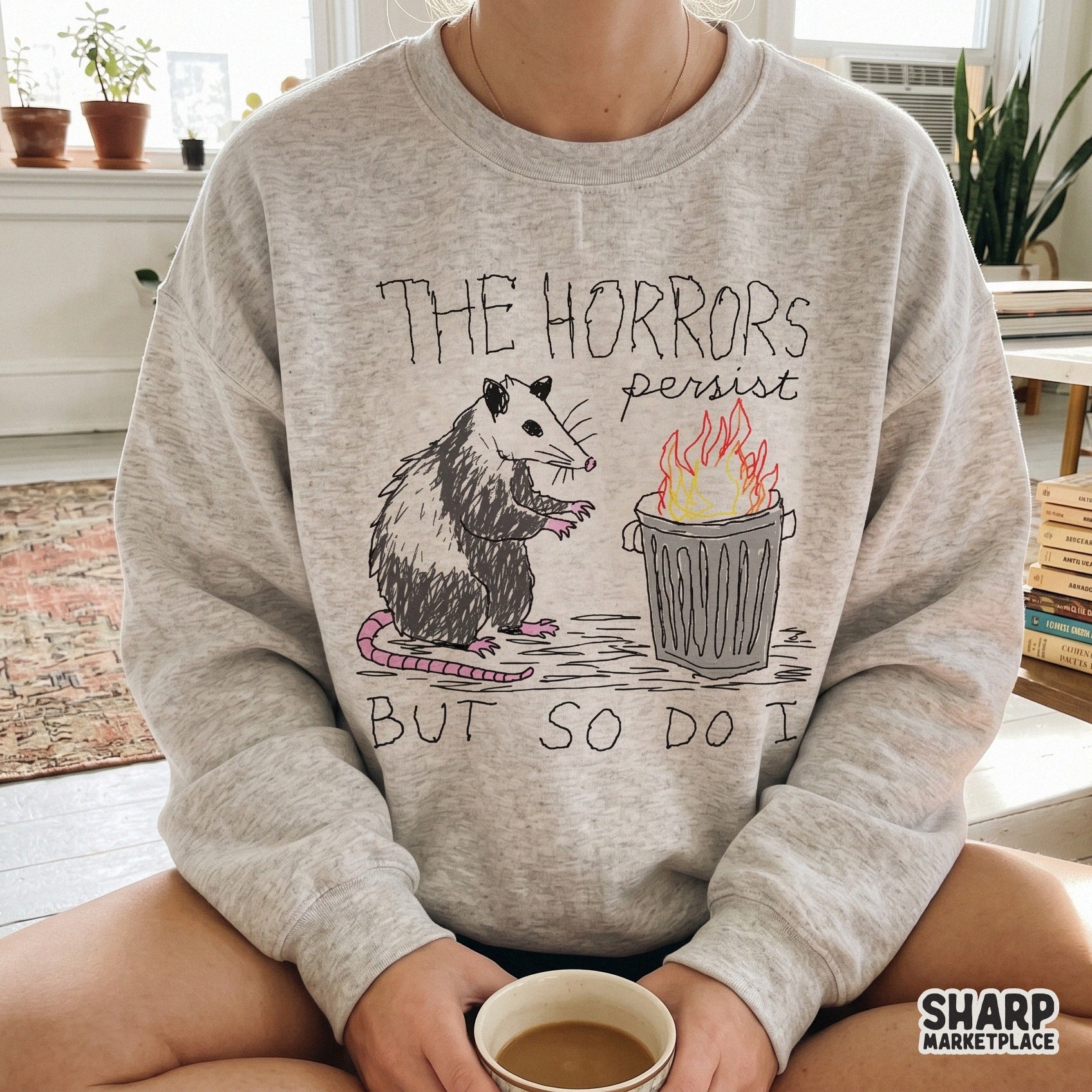 Funny opossum meme design on a grey sweatshirt showing a scribbled possum near a trash fire with the quote The Horrors Persist But So Do I.