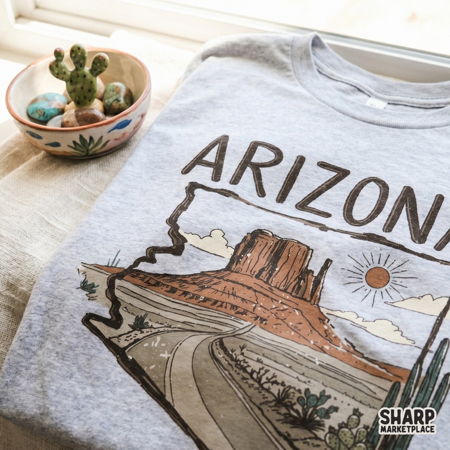 Southwest desert landscape illustration framed by the Arizona state outline, featuring a scenic highway, saguaro cacti, and a warm sun motif.