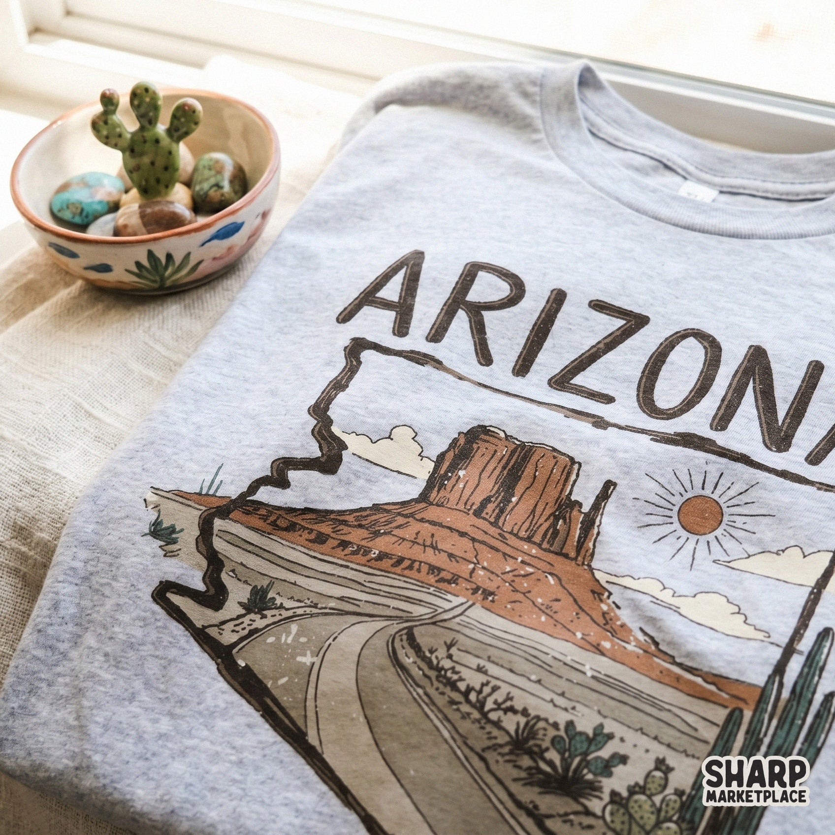 Southwest desert landscape illustration framed by the Arizona state outline, featuring a scenic highway, saguaro cacti, and a warm sun motif.