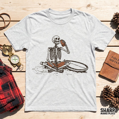 A grey t-shirt mockup featuring a retro skeleton design sitting cross-legged on a beach with a surfboard and a coconut drink.