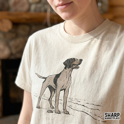 Detailed hand-drawn pointer dog illustration on a coastal shore, displayed as a graphic design on a light-colored t-shirt.