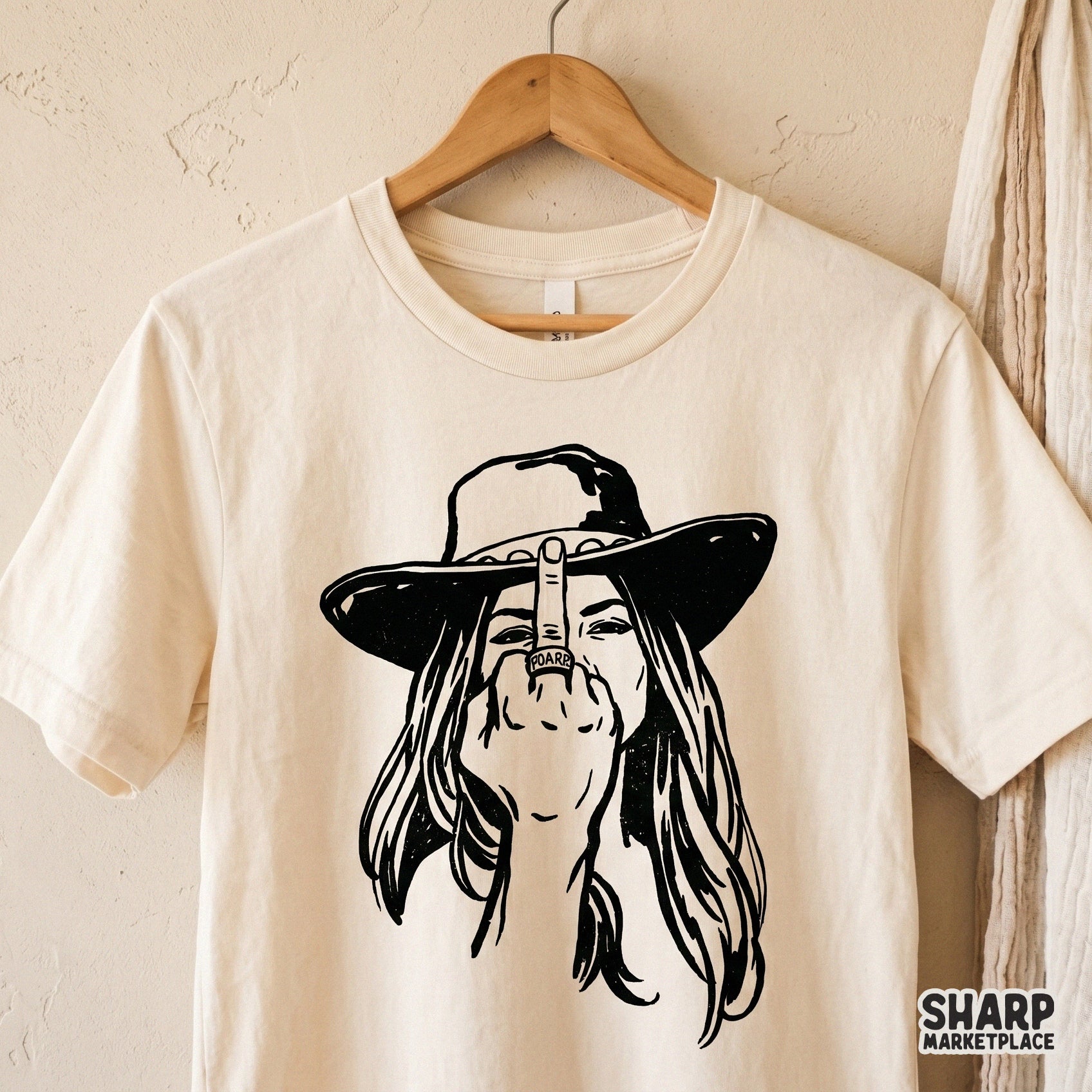 Mockup of a cream colored t-shirt featuring a black ink sketch of a sassy cowgirl wearing a cowboy hat and gesturing with a ring that says FUCK OFF.