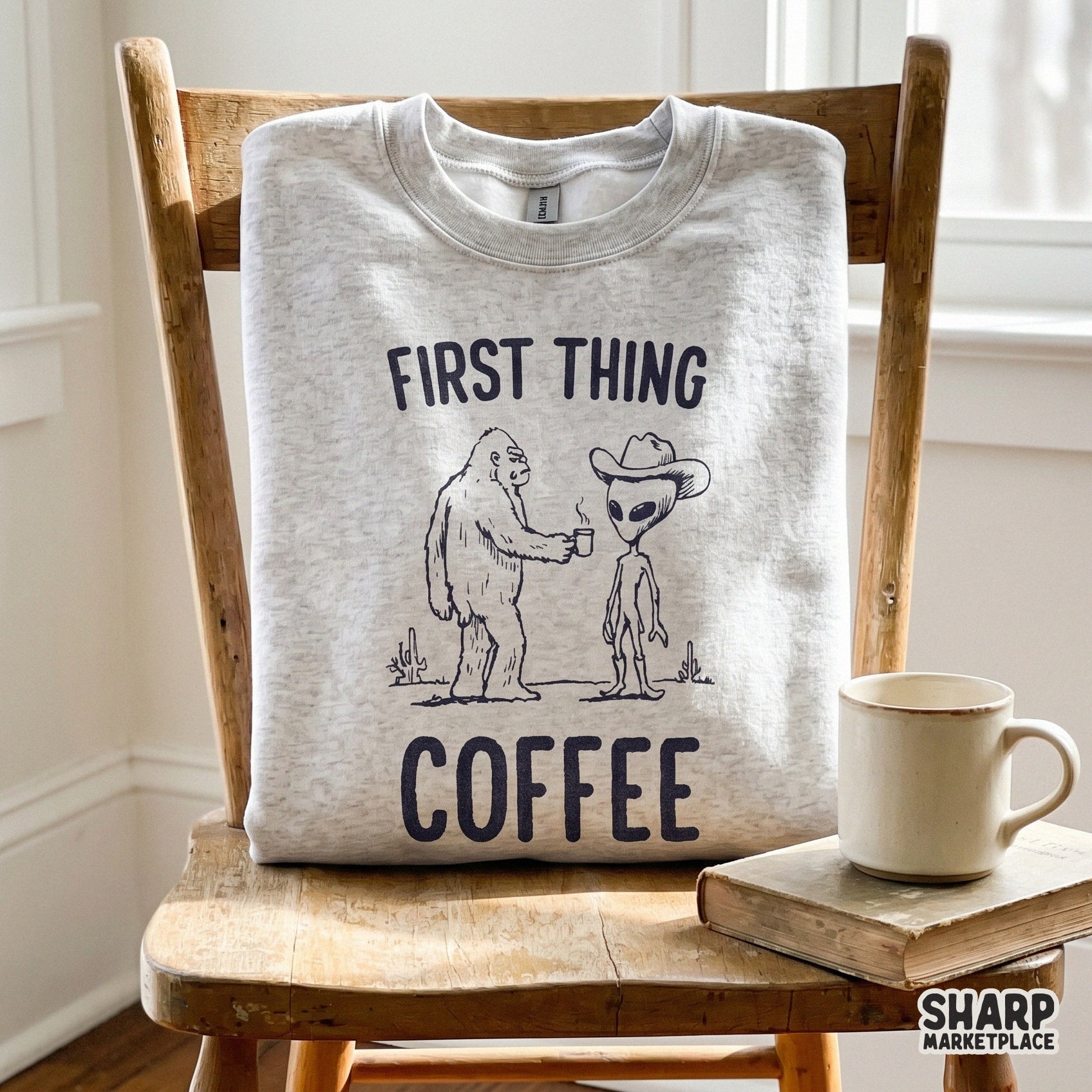 A light-colored sweatshirt folded on a wooden chair featuring a monochrome line art design of a cryptid and alien cowboy sharing coffee with desert motifs.