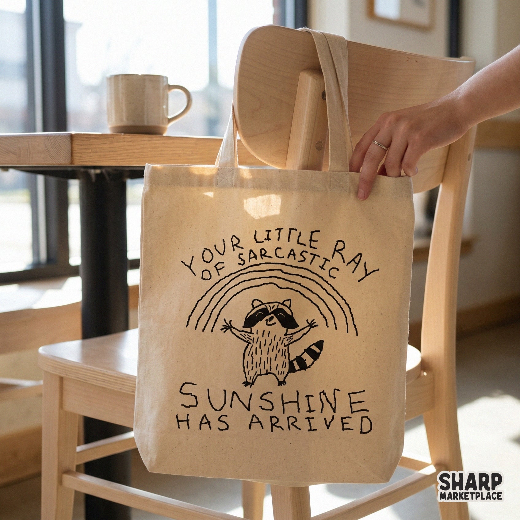A tote bag featuring a cute raccoon standing under a rainbow with the witty quote Your Little Ray Of Sarcastic Sunshine Has Arrived.