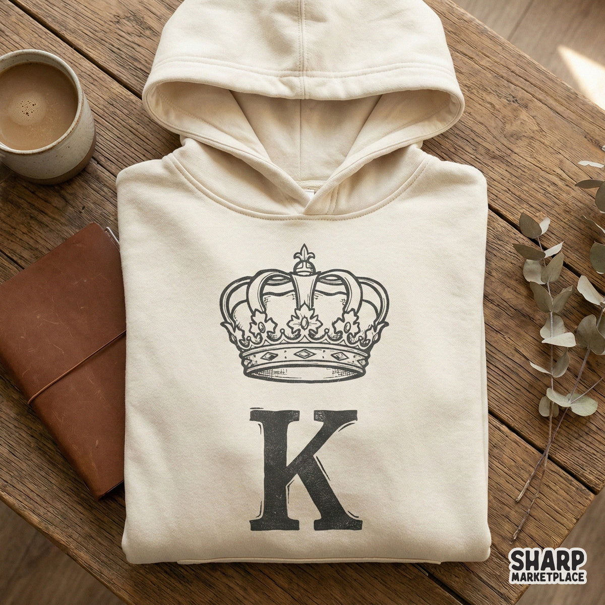 A beige hoodie mockup displaying a bold serif letter K topped with a regal crown, rendered in a sophisticated sketch style for personalized clothing.