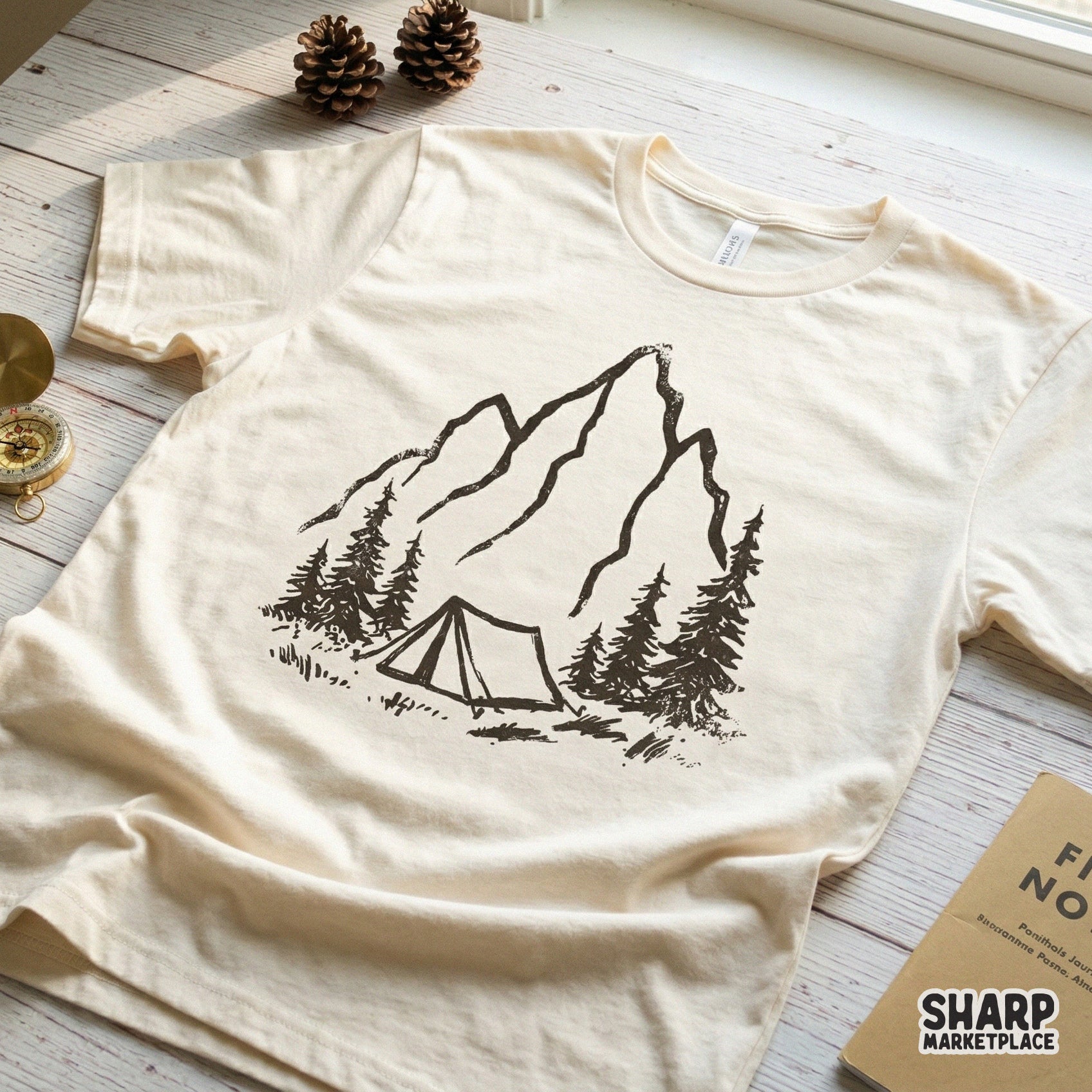 A textured black sketch of a mountain summit and camping tent surrounded by pine trees, featured as a trendy graphic on a cream-colored t-shirt.