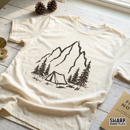 A textured black sketch of a mountain summit and camping tent surrounded by pine trees, featured as a trendy graphic on a cream-colored t-shirt.