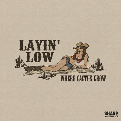 Vintage western digital design featuring a cowgirl in a hat and denim shorts lounging on a rug with cacti, text reads Layin Low Where Cactus Grow.