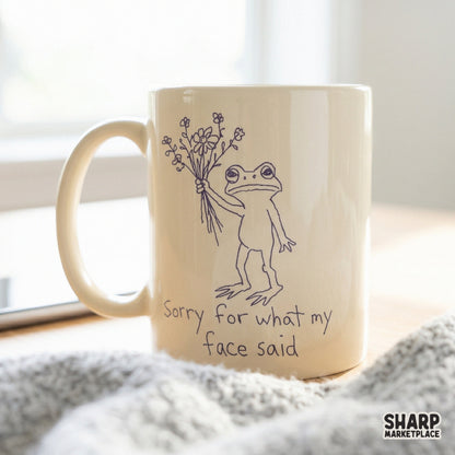 A minimalist digital design featuring a deadpan frog with flowers and the quote Sorry for what my face said, displayed on a cream-colored coffee mug.