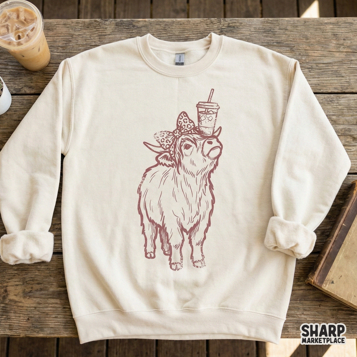 Rustic monochromatic Highland cow graphic with a leopard bow and iced coffee, shown as a digital design on a neutral-colored crewneck sweatshirt.