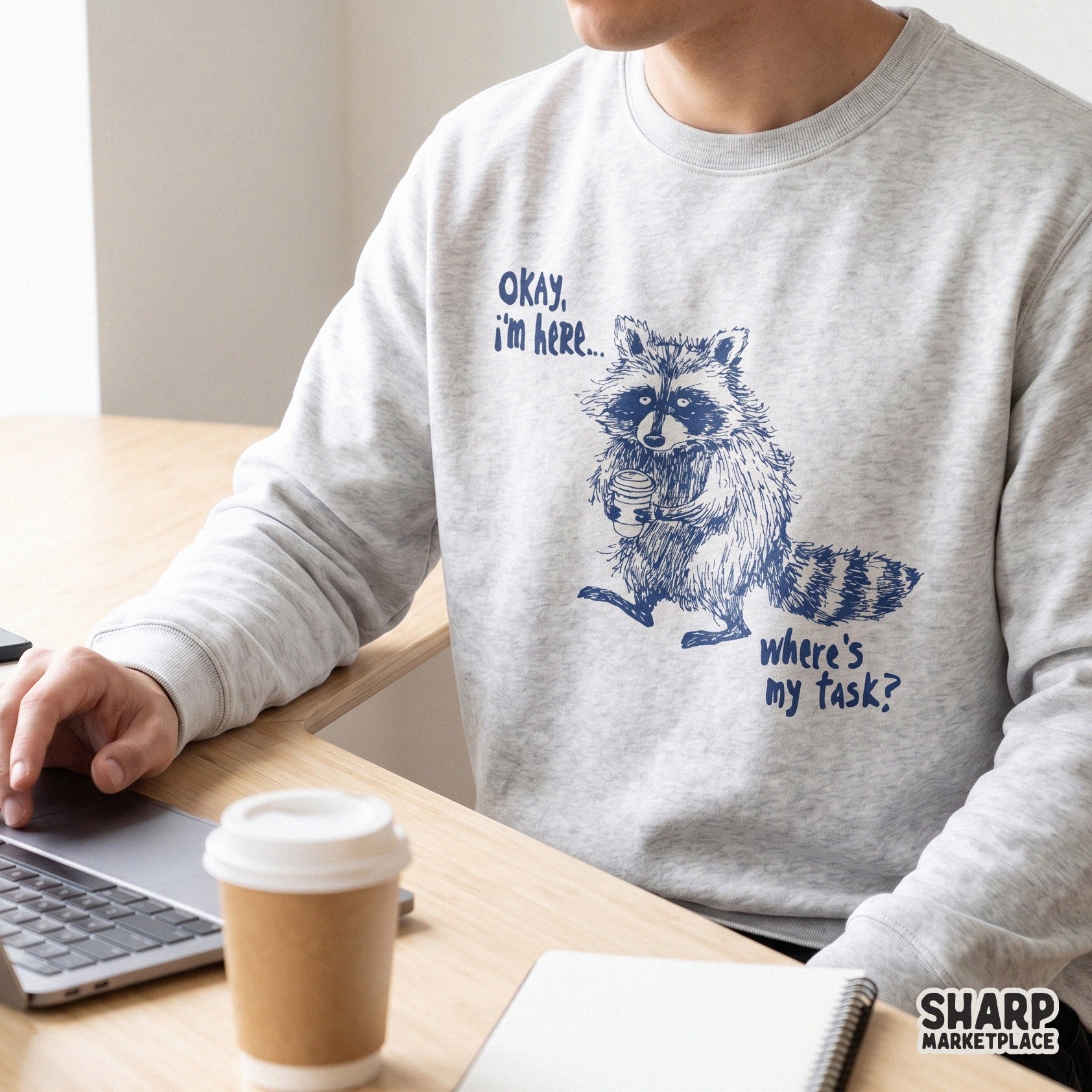 A light gray sweatshirt featuring a navy blue graphic of a raccoon clutching a coffee cup with the sarcastic caption Okay, I'm Here... Where's My Task?