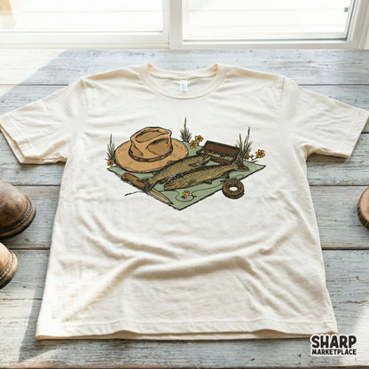 Rustic fly fishing illustration on a cream t-shirt mockup, showcasing trout, fishing gear, and wildflowers in a vintage hand-drawn art style.