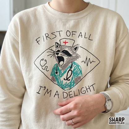 Sarcastic healthcare digital download featuring a raccoon in a nurse cap and scrubs with the text First Of All I'm A Delight for DIY shirts and tumblers.