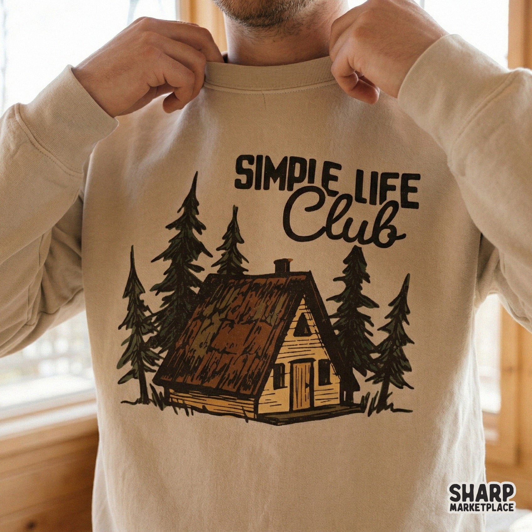 Trendy sublimation design on a beige sweatshirt showing a rustic A-frame cabin nestled in a pine forest with the text Simple Life Club in earthy tones.