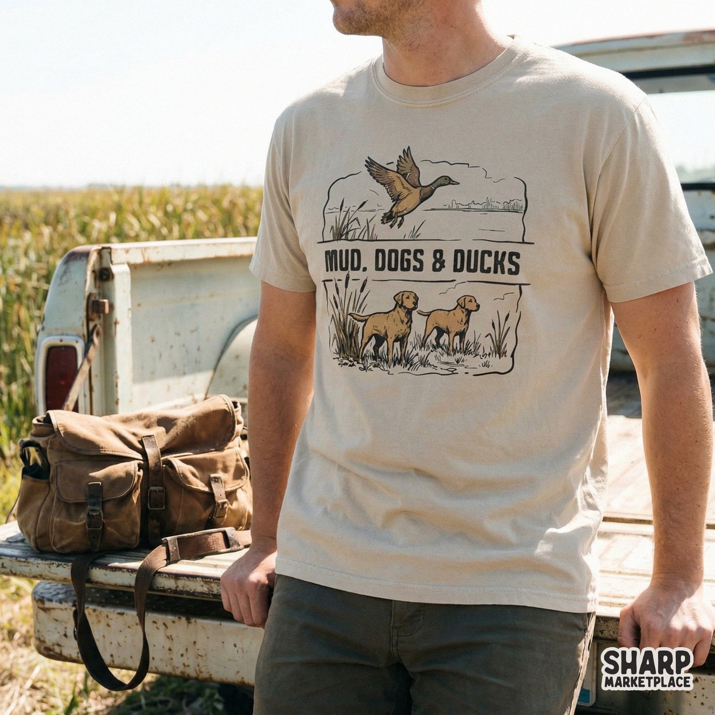 Rustic outdoor digital illustration showing a flying mallard and two dogs in cattails, displayed as a graphic print on a tan t-shirt.