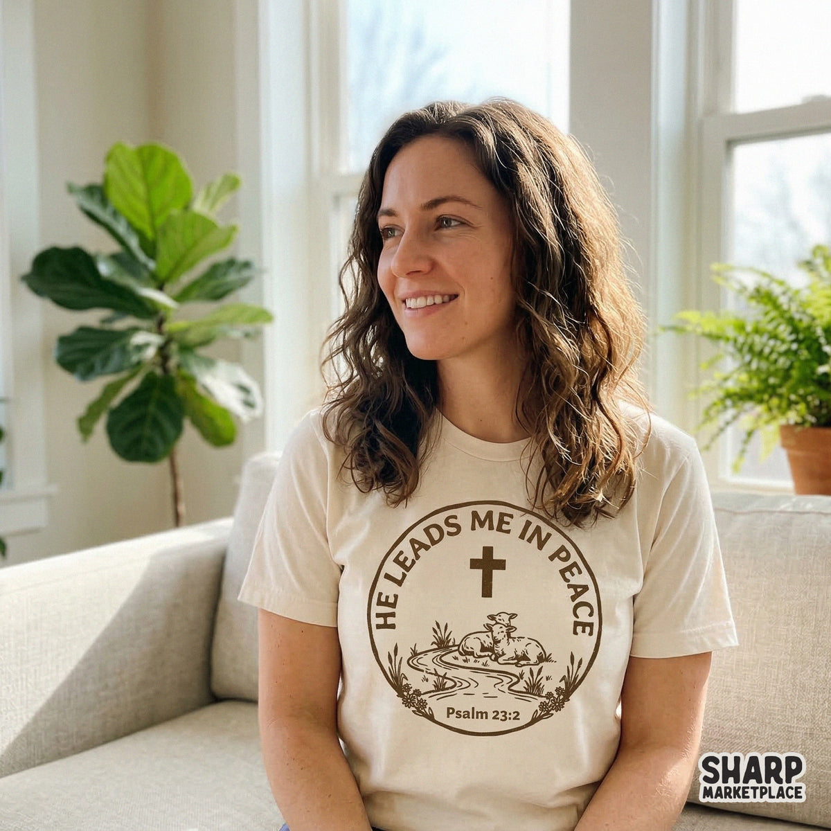 A circular Christian design with two resting lambs and a cross, featuring the scripture He Leads Me In Peace, shown on a cream-colored t-shirt.