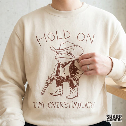 Transparent PNG file of a doodle-style cowboy frog wearing a tiny holster and cowboy hat, paired with the text Hold On I'm Overstimulated for print-on-demand.