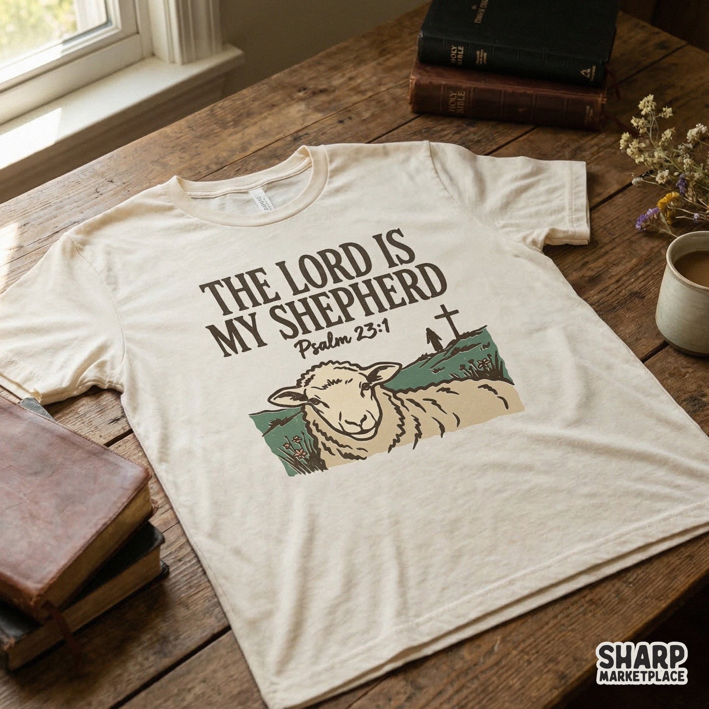 Cream colored t-shirt mockup displaying a rustic Bible verse design with a hand-drawn sheep illustration and a distant cross on a hill.