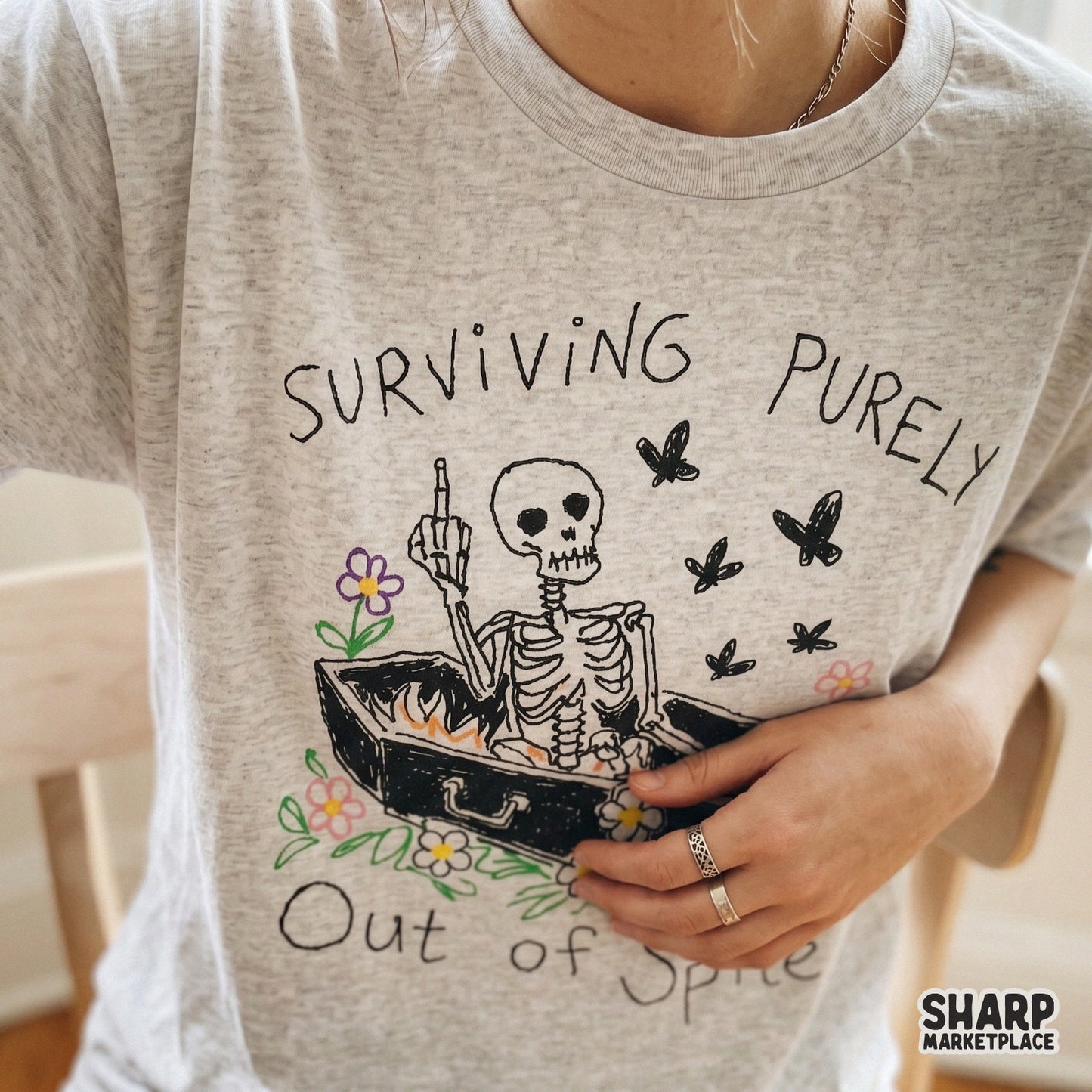 Trendy apparel mockup featuring a skeleton lounging in a coffin with colorful floral accents and the text Surviving Purely Out Of Spite on a heather shirt.