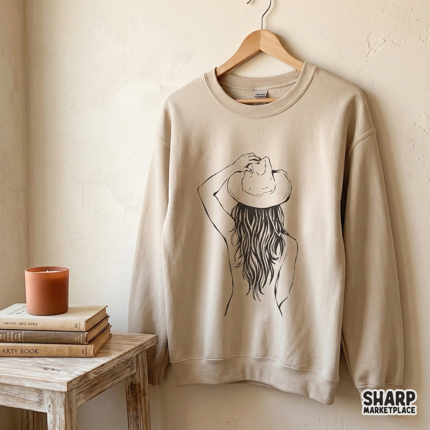 A beige crewneck sweatshirt featuring a black ink line art illustration of a cowgirl with long wavy hair and a western hat.