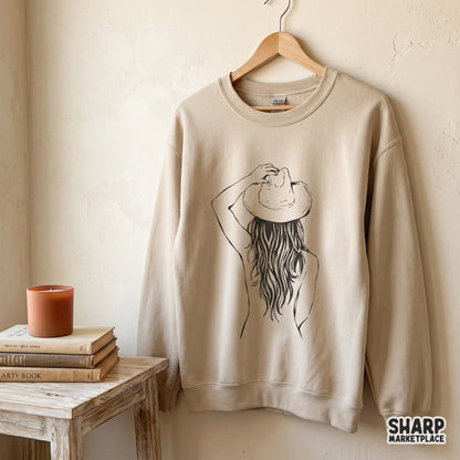 A beige crewneck sweatshirt featuring a black ink line art illustration of a cowgirl with long wavy hair and a western hat.