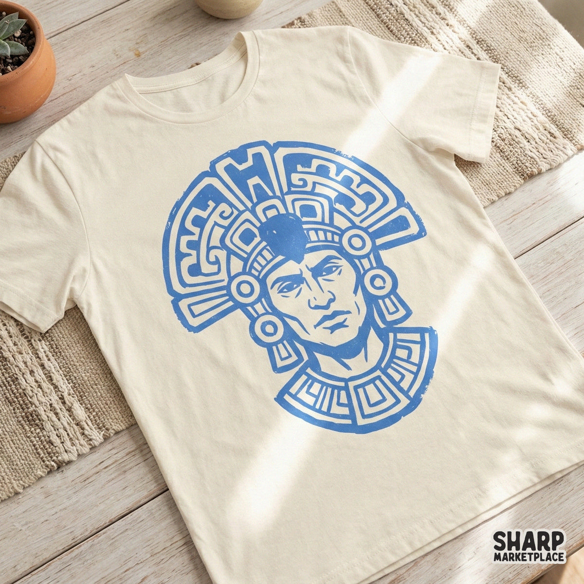 A vintage-style blue Aztec warrior graphic with a detailed headdress and collar, displayed on a cream-colored t-shirt for apparel design inspiration.