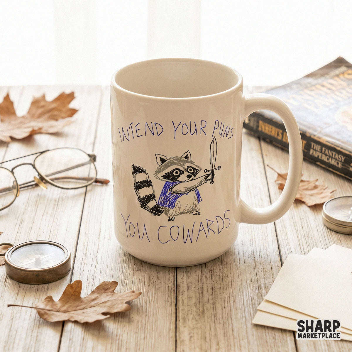 A white ceramic coffee mug on a wooden table featuring a whimsical sketch of a sword-wielding raccoon in a blue cape with the text Intend Your Puns You Cowards.