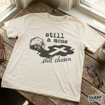 Still a mess still chosen Christian design printed on a cream-colored t-shirt, showcasing a spilled ink bottle and a cross silhouette for faith-based apparel.