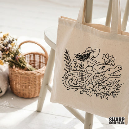 Whimsical digital illustration of a woman riding an alligator while holding flowers, shown as a black design on a canvas tote bag.