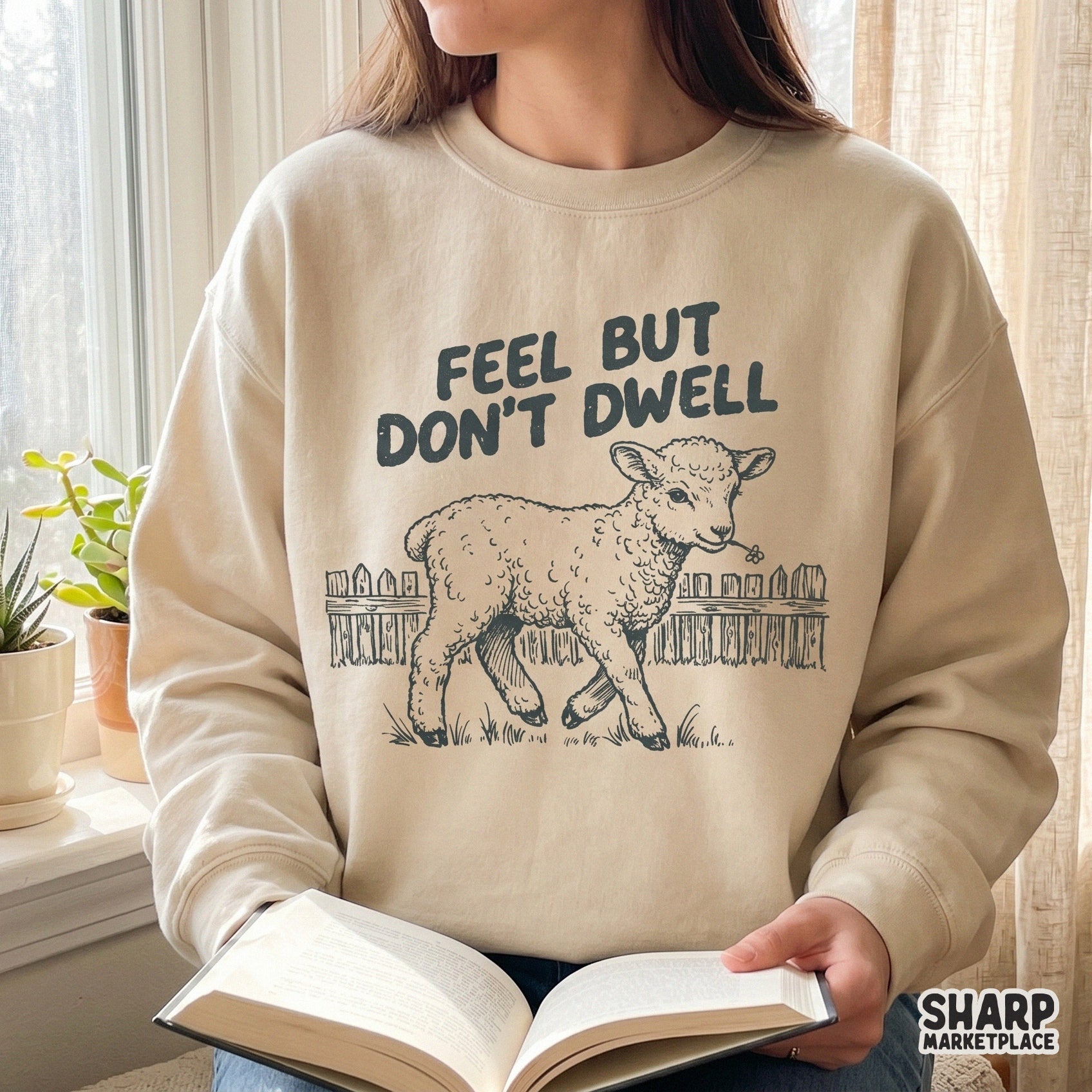 A lifestyle mockup showing a beige crewneck sweatshirt featuring a hand-drawn lamb illustration and the inspirational quote Feel But Don't Dwell.