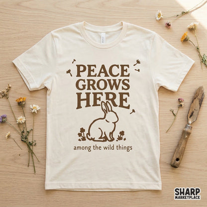 A cream-colored t-shirt mockup displaying a brown graphic of a bunny in a field of wildflowers with the inspirational text Peace Grows Here.
